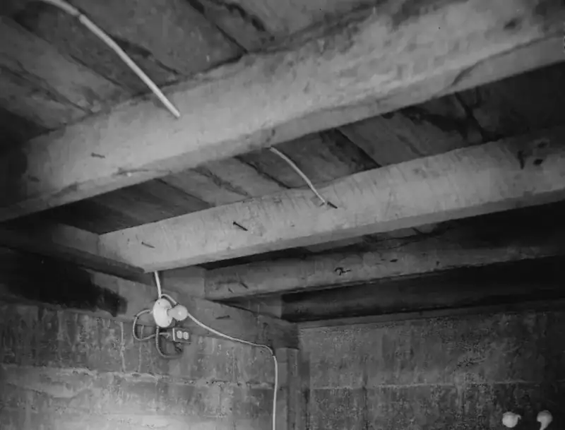 Black and white photo of an unfinished basement corner with rough board-formed concrete walls and heavy timber floor beams overhead