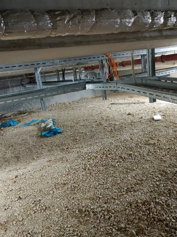 Crawl space under a building with insulated ductwork overhead, a red drain pipe, metal cable trays carrying wiring, a gravel floor and discarded plastic and gloves on the ground