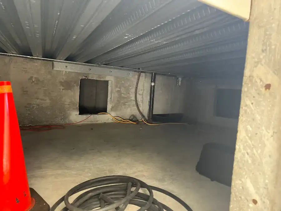Crawl space with a concrete floor, concrete walls, metal deck overhead, an opening in the far wall, extension cords and a traffic cone near the entrance