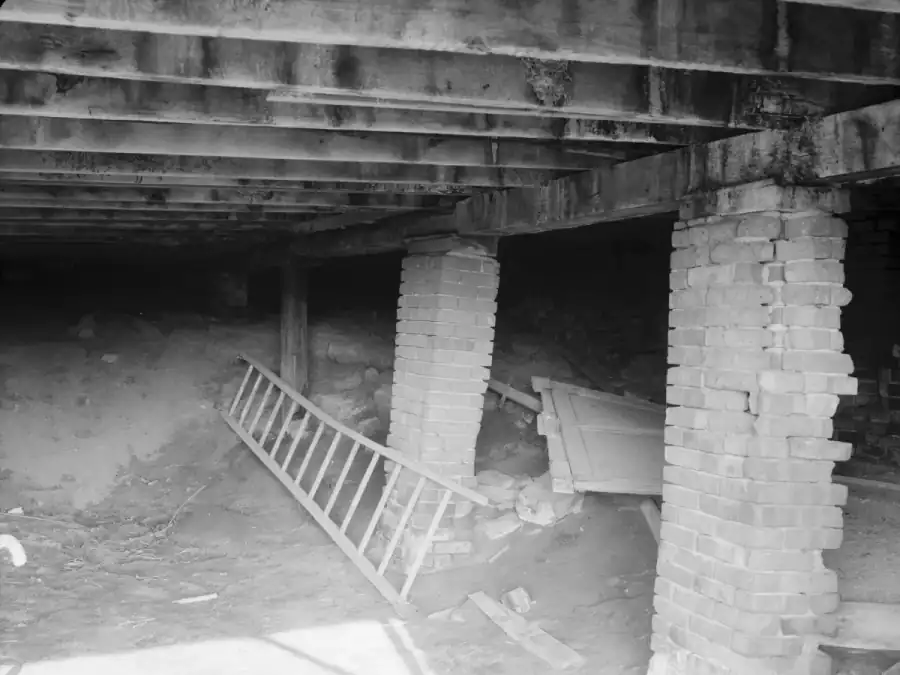 Old crawl space with two brick piers that are bulging and losing bricks under timber beams and joists, with a wooden post, a ladder and debris on the dirt floor