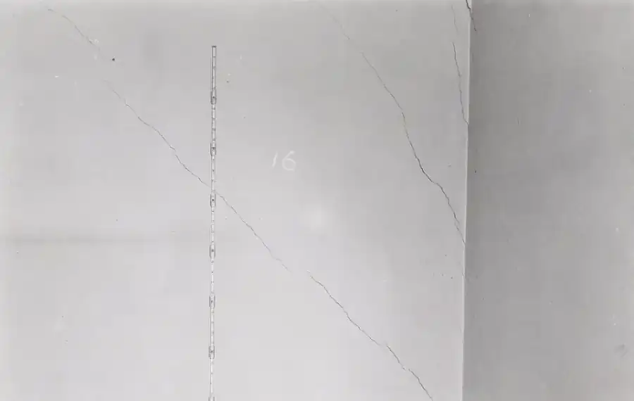 Black and white photo of a plaster bedroom wall with two long diagonal cracks running down toward a corner and a measuring rod held against the wall for scale