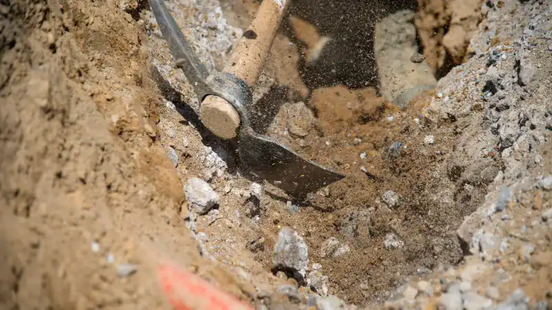 Pickaxe striking the rocky soil at the bottom of a narrow trench dug for a French drain