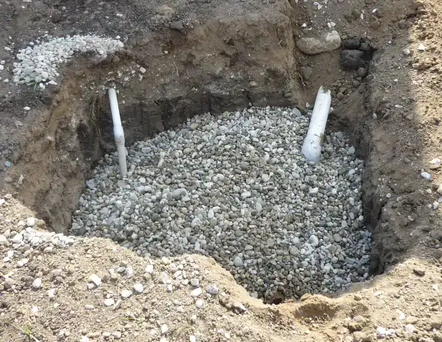 Excavated pit in bare soil filled with washed gravel, with two white plastic pipes rising out of the stone