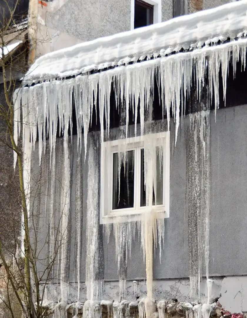 Thick snow on a roof edge with a row of long icicles hanging over a window and running down the wall below, a sign of an ice dam
