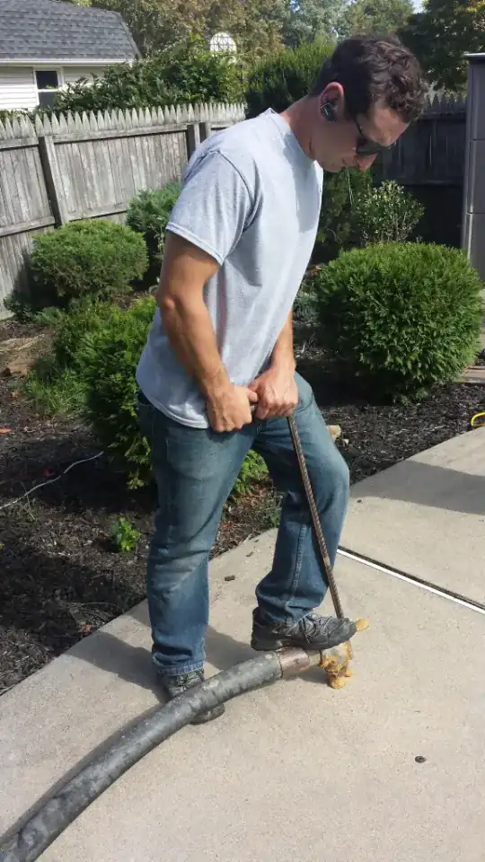 Worker holding a mudjacking hose nozzle in a hole drilled through a concrete patio slab while slurry is pumped underneath
