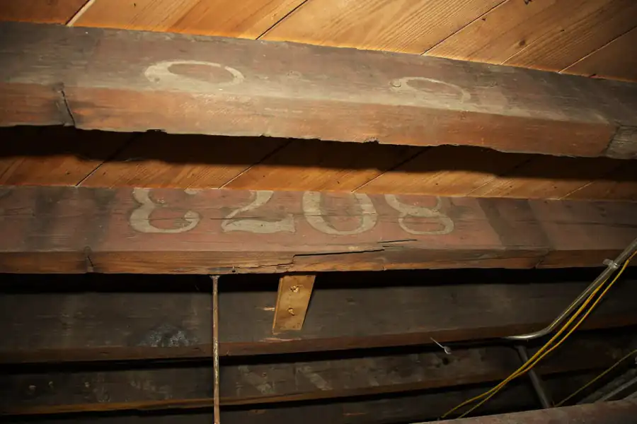 Underside of old wooden floor joists in a basement, with stenciled numbers on the timbers and a long split running along the bottom edge of one joist