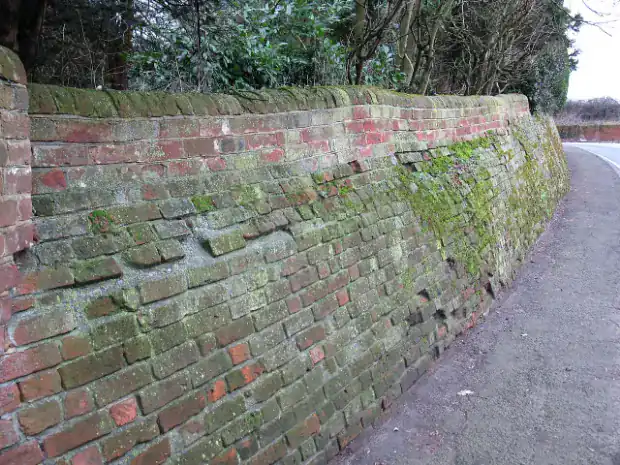 Old red brick wall holding back a raised bank, bulging outward along its length with cracked and shifted mortar courses near mid-height