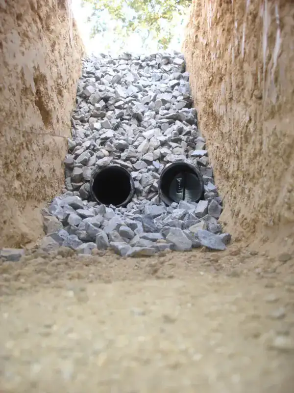 Narrow trench dug in sandy soil with two black perforated drain pipes laid on a bed of gravel and more gravel heaped over them