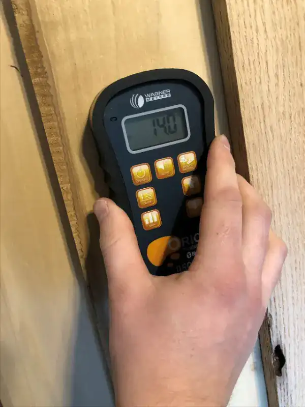 Hand holding a pinless wood moisture meter flat against a board, with the display showing a moisture content of 14 percent