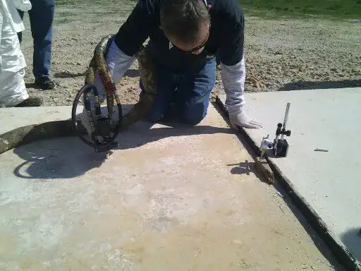 Technician kneeling on a concrete slab with a polyurethane foam injection gun and hose, with a dial gauge set up at the slab edge to measure the lift