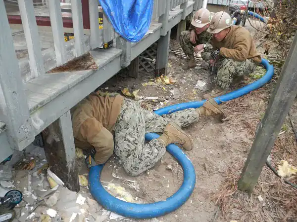 Navy construction crew members kneeling beside a raised house, one reaching under the floor with a large blue suction hose to pump out floodwater, with soaked insulation scattered on the ground