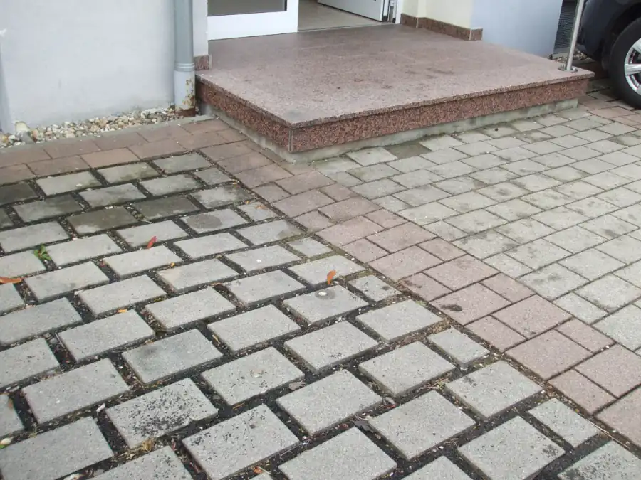 Paving beside a house entrance that has dropped several centimeters below a granite step, exposing the step's concrete base where backfill soil settled