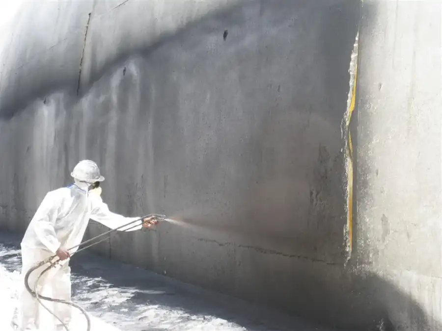 Worker in a white protective suit and hard hat spraying a dark liquid waterproofing membrane onto the outside face of a tall concrete wall