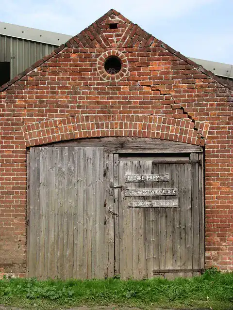Red brick gable wall with a stair-step crack running through the mortar joints from the eaves down to the brick arch over a pair of wooden doors