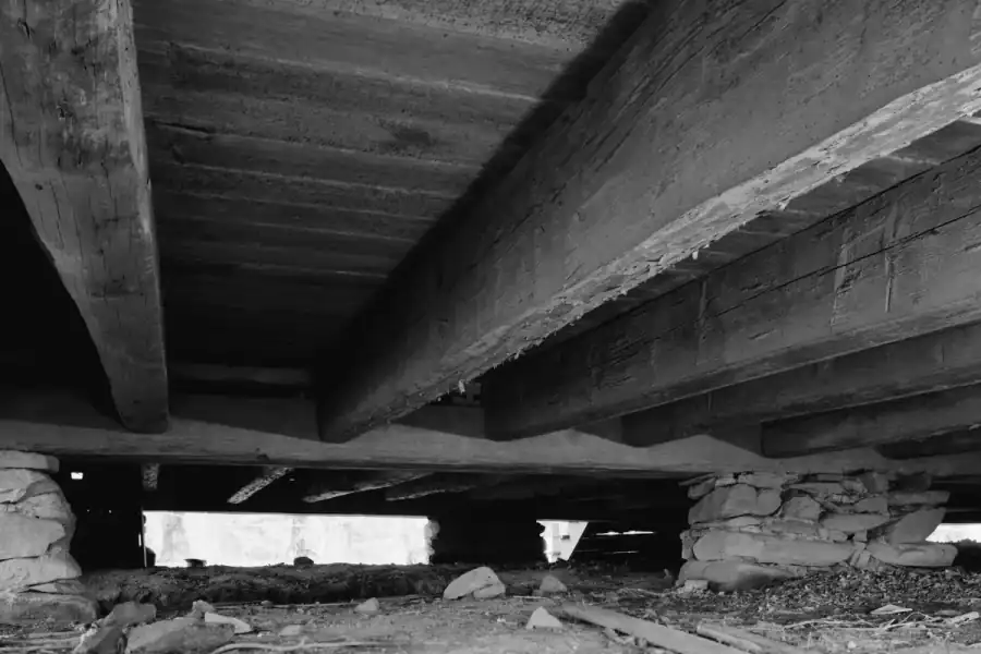 Crawl space under a historic house showing dry-stacked stone piers on bare soil carrying hand-hewn timber beams and floor joists