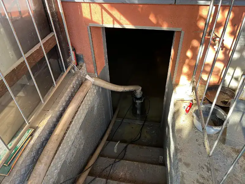 Submersible electric pump standing in floodwater at the bottom of an outside stairwell to a flooded lower level, with a discharge hose running up the steps