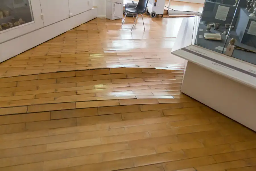 Wood strip floor in a room with rows of boards swollen, raised and pushed out of line after a water leak, with drying equipment running in the background