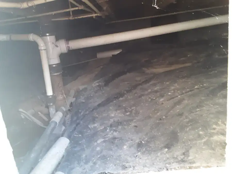 Crawl space seen through an access opening with an old, dirty plastic vapor barrier partly covering the soil and plumbing pipes running overhead