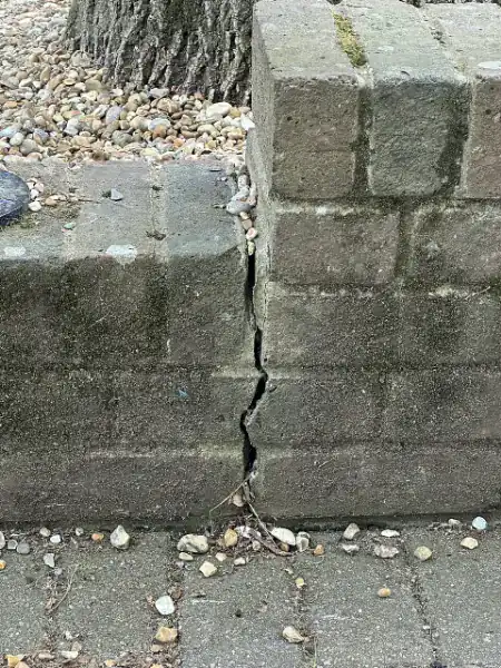 Concrete block retaining wall with a wide vertical crack running through the blocks from top to bottom, gravel on the soil side above