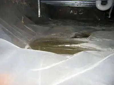 Muddy water pooled on top of clear plastic sheeting on a crawl space floor, under plumbing pipes